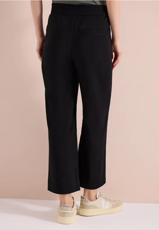 Achteraanzicht van een vrouw in zwarte, wijdvallende, enkellange broek en beige sneakers.