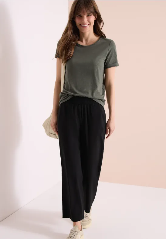 Vrouw in casual outfit met groen T-shirt, zwarte wijde broek en sneakers voor een lichte achtergrond.