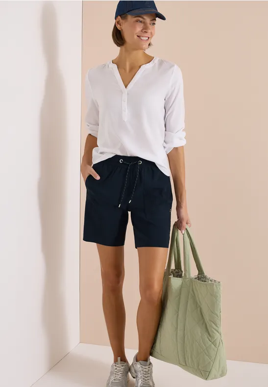 Iemand met witte blouse, donkerblauwe short, grijze sneakers en een grote groene tas.