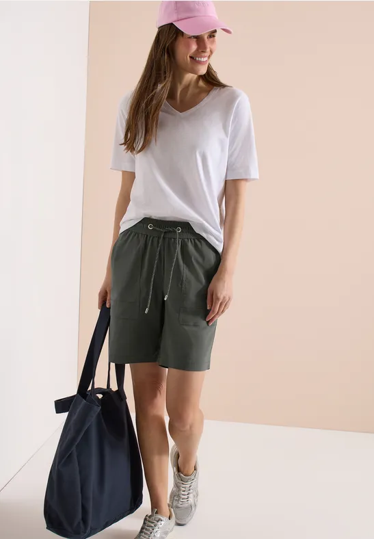 Jonge vrouw in wit T-shirt, kaki shorts, roze pet en sneakers, draagt een donkerblauwe tas.