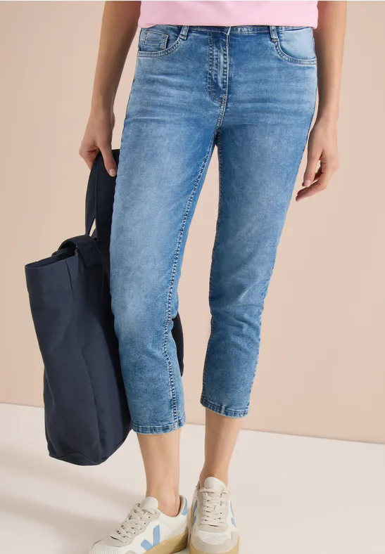 Frau trägt blaue Cropped-Jeans und weiße Sneaker, hält dunkelblaue Tasche vor beigem Hintergrund.