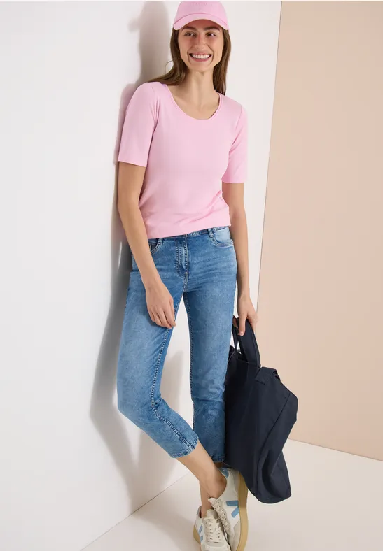 Person lehnt an weißer Wand, trägt rosafarbenes T-Shirt, blaue Jeans und hält eine dunkle Jacke.