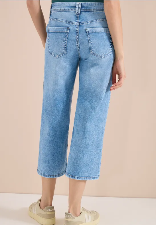 Achteraanzicht van een vrouw in wijdvallende, korte spijkerbroek en lichte sneakers voor een beige achtergrond.