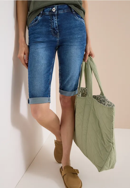 Person trägt blaue Denim-Bermudashorts, olivgrüne Tasche und braune Slipper vor neutralem Hintergrund.
