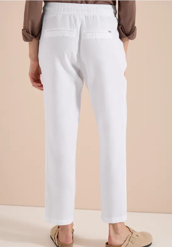 Rückansicht einer Person in weißen Leinenhosen und beigen Loafers vor hellem Hintergrund.