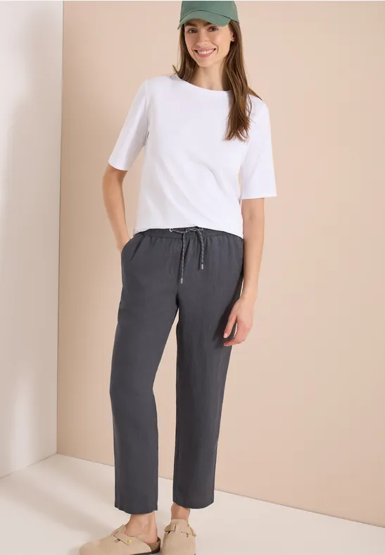 Frau trägt weißes T-Shirt und dunkelgraue, lockere Hose vor beigem Hintergrund.