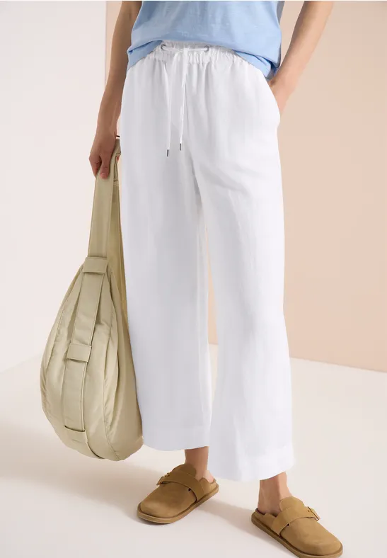Weiße Leinenhose mit Kordelzug, beige Slipper und große Tasche vor hellem Hintergrund.