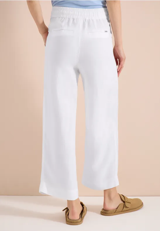 Rückansicht einer Frau in weißen, weit geschnittenen Leinenhosen und Mokassins vor beigem Hintergrund.
