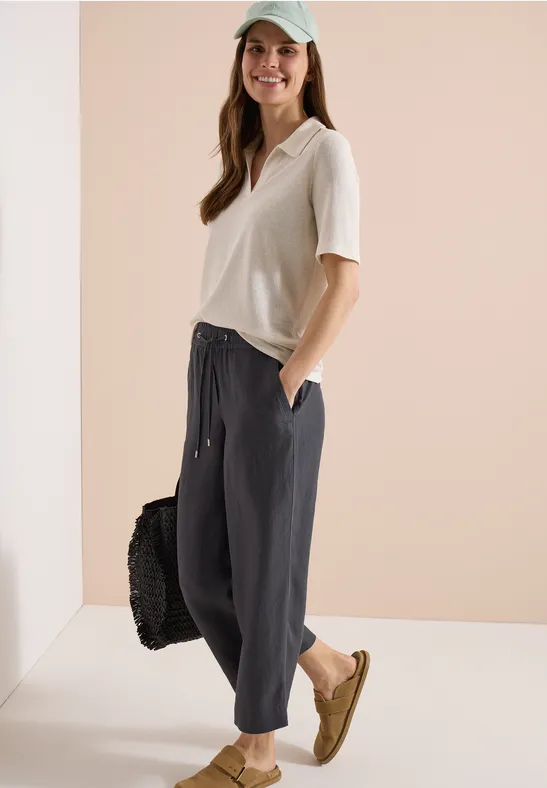 Frau trägt lockeres weißes T-Shirt, weite schwarze Hose, beige Schuhe und hält eine schwarze Tasche.