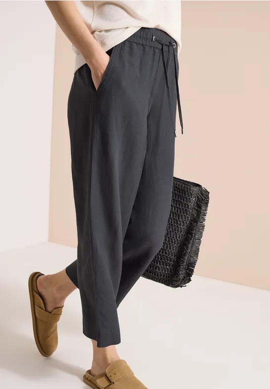 Schwarze, locker sitzende Leinenhose mit Kordelzug, kombiniert mit senfgelben Slippers und einer schwarzen Korbtasche.