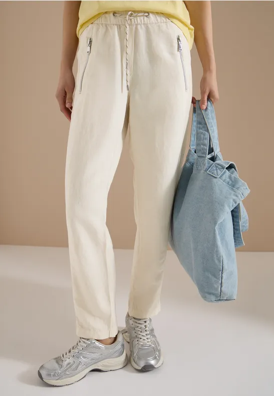 Person trägt weiße Hosen, graue Sneaker und hält eine hellblaue Tasche.