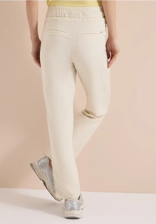 Rückansicht einer Person in hellen Jogginghosen und silbernen Sneakern vor neutralem Hintergrund.