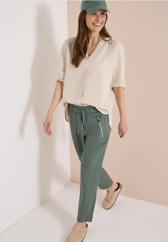 Frau in beiger Leinenbluse, grünen Jogginghosen und Sneakers vor hellem Hintergrund.