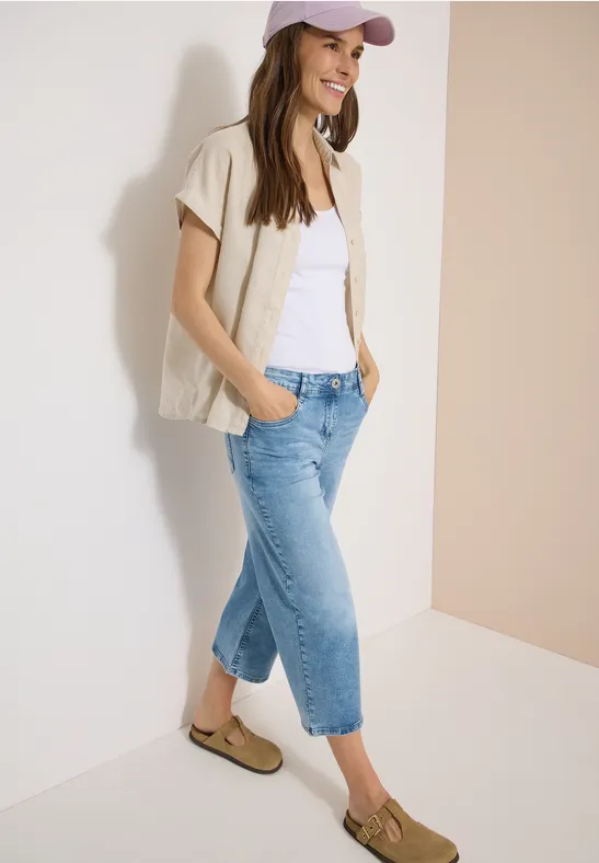 Junge Frau lehnt lässig an Wand, trägt beiges Kurzarmhemd, weißes Top, blaue Jeans, Loafer und Cap.