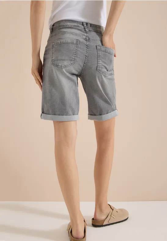 Rückansicht eines Mannes in grauen Jeans-Shorts mit Umschlag und beigen Mokassins vor hellem Hintergrund.