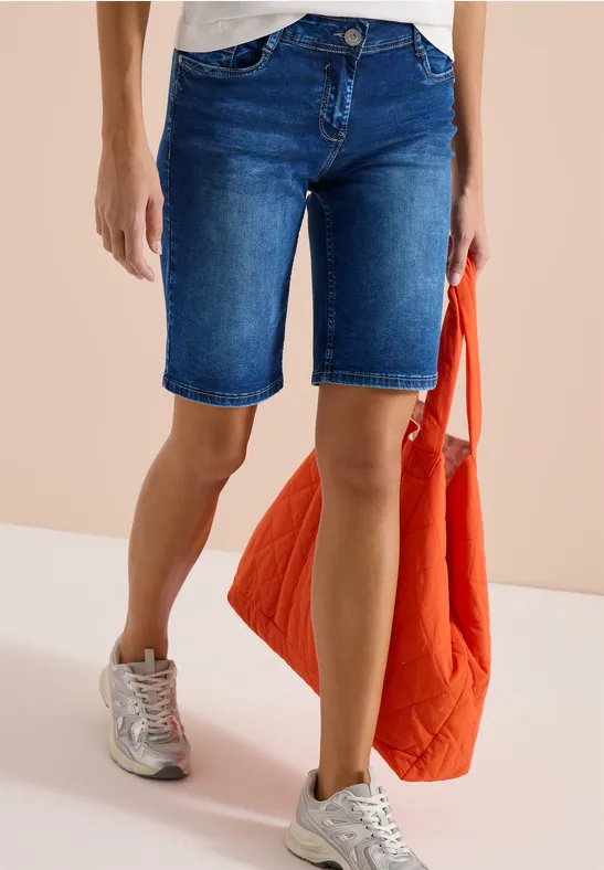 Frau trägt knielange Jeans-Shorts, weiße Sneaker und hält eine gesteppt orange Jacke in der Hand.