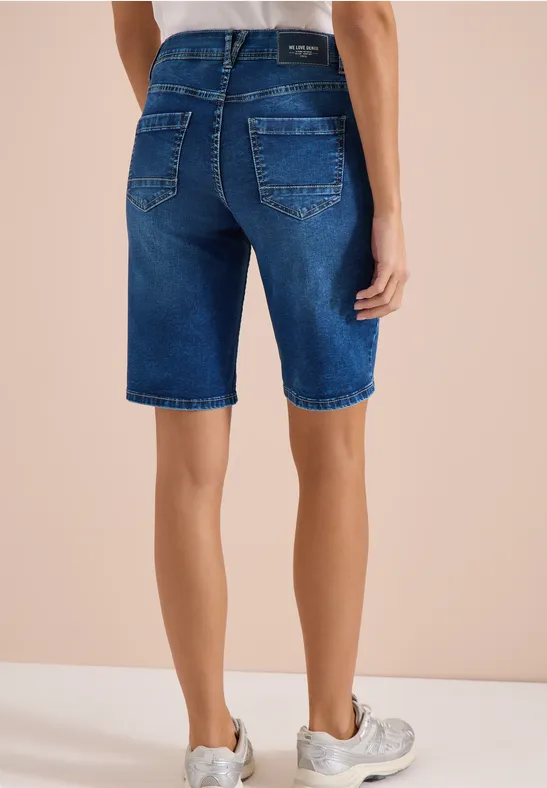 Rückansicht einer Person in dunkelblauen Jeans-Bermudas und weißen Sneakern vor beigem Hintergrund.