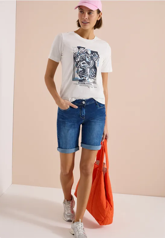 Frau in weißem T-Shirt mit Grafik, Jeans-Shorts, Sneakern und roter Tasche.