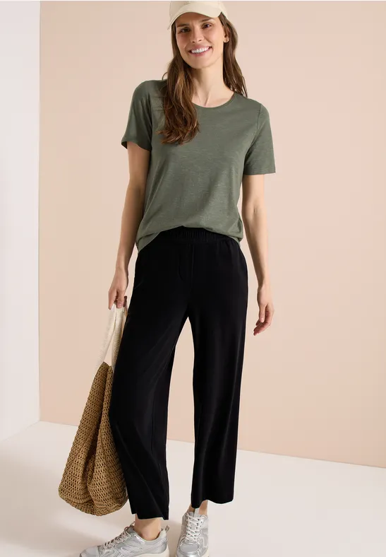 Frau trägt grünes T-Shirt, weite schwarze Hose, weiße Sneaker und Korbtasche vor neutralem Hintergrund.