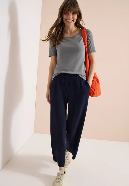 Eine Frau trägt graues T-Shirt, dunkelblaue Hose, weiße Sneaker und eine orangefarbene Umhängetasche.