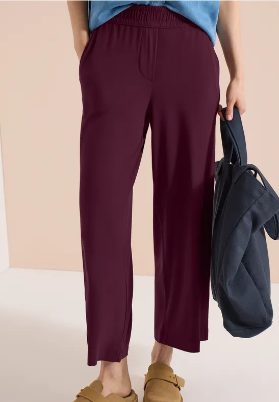 Eine Person trägt eine weite, bordeauxrote Hose, hellblaues Oberteil, Sandalen und hält eine dunkelblaue Tasche.