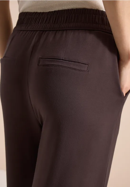 Nahaufnahme: Person trägt dunkelbraune Hose mit elastischem Bund, Seitentaschen und Paspeltasche.