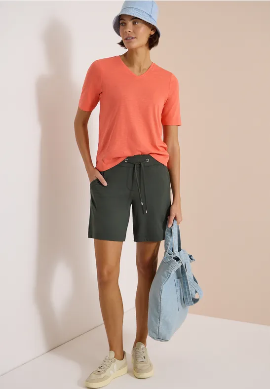 Person trägt korallenfarbenes T-Shirt, dunkelgrüne Shorts, Sneaker und hält eine Jeansjacke.