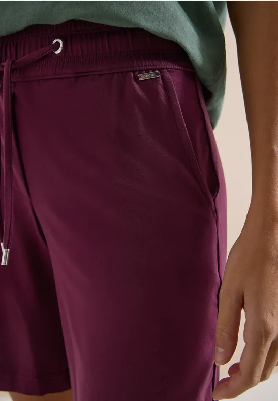 Nahaufnahme einer Person in weinroten Shorts und grünem Shirt, mit Tasche und Kordelzug.