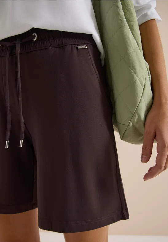 Nahaufnahme: Eine Person trägt dunkelbraune Shorts, ein weißes Shirt und eine grüne, gesteppte Umhängetasche.