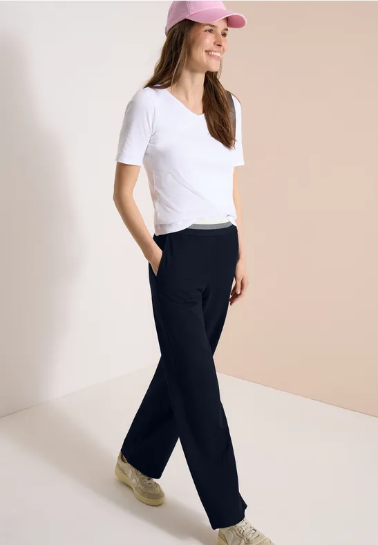 Femme en t-shirt blanc, pantalon noir décontracté, baskets et casquette rose sur fond clair.