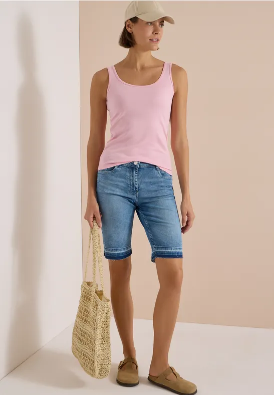 Femme portant débardeur rose, bermuda en jean, casquette, sac en paille et mocassins devant un mur beige.