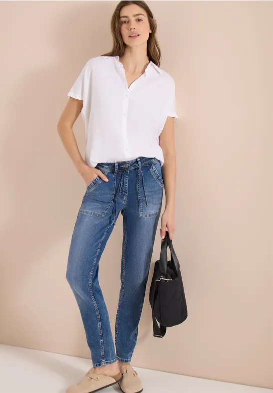 Frau in weißem Poloshirt, blauen Jeans mit Taillengürtel und Espadrilles trägt eine schwarze Tasche.