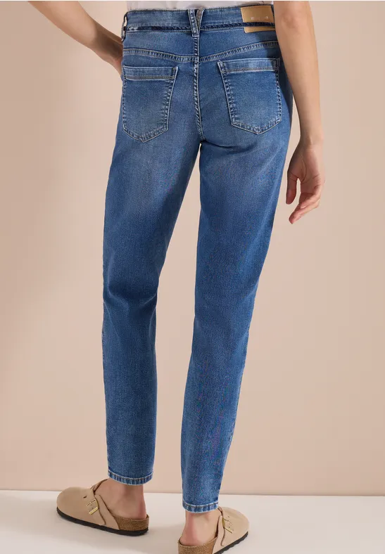 Person von hinten in blauen, leicht taillierten Jeans und beigen Pantoletten vor hellem Hintergrund