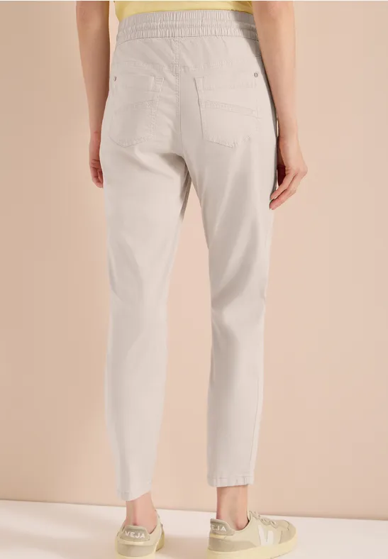 Rückansicht einer Frau in locker sitzender, hellgrauer, hoch taillierter Hose vor beigem Hintergrund.