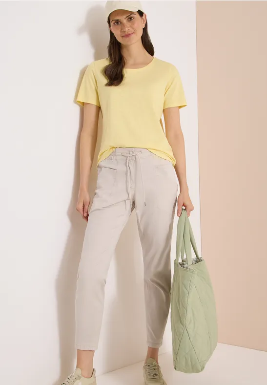 Frau in gelbem T-Shirt, hellgrauen Hosen und grünem Stepp-Shopper vor schlichtem, pastellfarbenem Hintergrund.