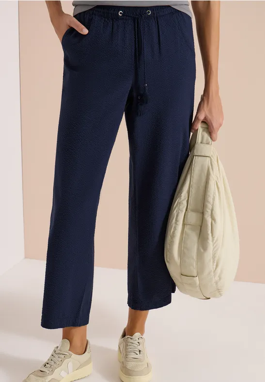 Loszittende marineblauwe broek met aantrekkoord, gecombineerd met lichte sneakers en een lichte jas in de hand.