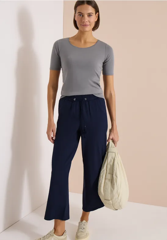 Vrouw met grijs shirt, wijde donkerblauwe broek en tas voor een neutrale achtergrond.