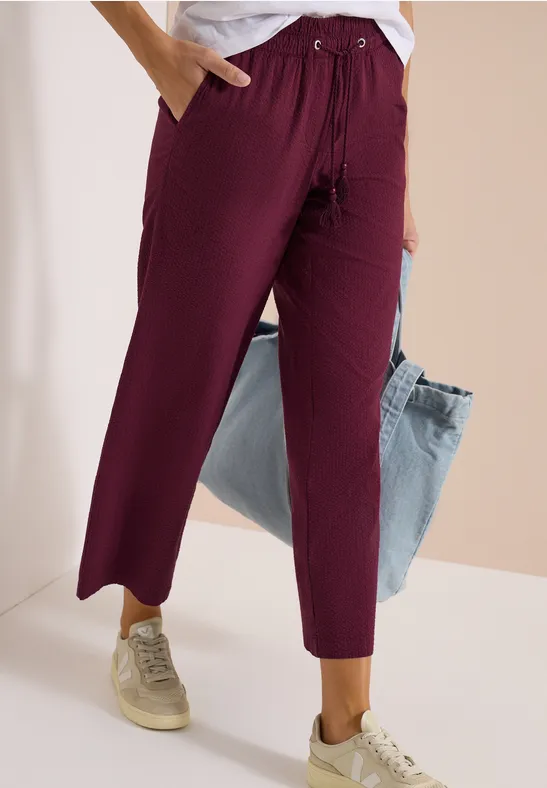 Slim-fit-Hose in dunklem Lila mit Tunnelzug und Taschen, kombiniert mit Sneakers und einer hellblauen Jacke.