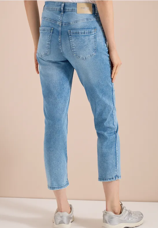 Vrouw in lichtblauwe, enkellange, rechte jeans met grijze sneakers tegen neutrale achtergrond.