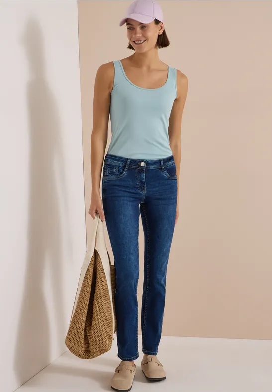 Jeune femme en jean, débardeur et casquette, portant un sac tressé.