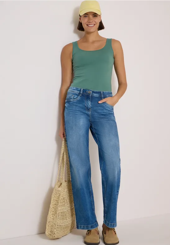 Frau mit gelber Cap, grünem Top, blauen Jeans und Strohtasche lächelnd