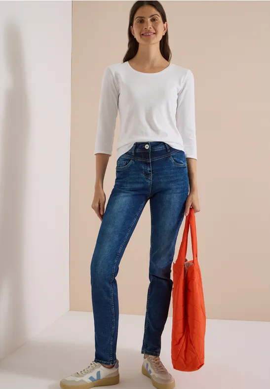 Frau in weißem Shirt und blauer Jeans, hält eine orangefarbene Tasche.