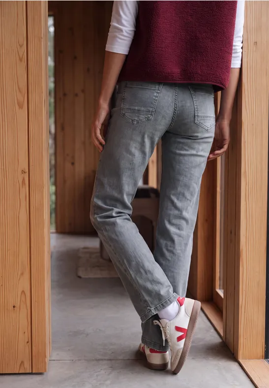 Person von hinten in grauen Jeans und weißen Sneakers steht in einer hölzernen Türöffnung.