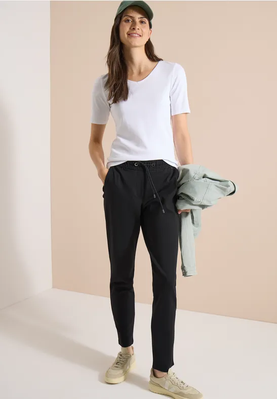 Frau in weißem T-Shirt und schwarzer Hose hält eine Jacke, minimalistische Umgebung, Sneaker an den Füßen.