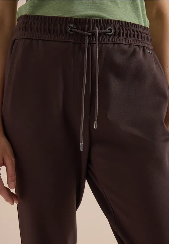 Person trägt dunkelbraune Jogginghose mit Tunnelzug und grünem Oberteil, Close-up auf Taille.