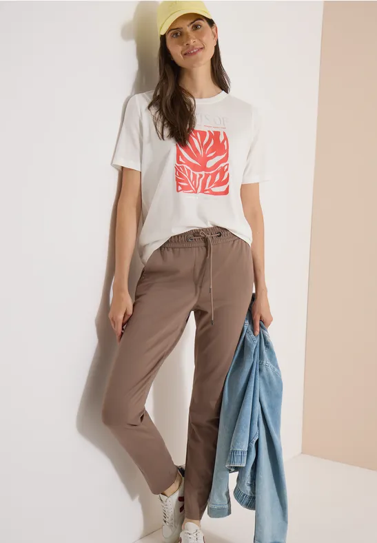 Frau trägt weißes T-Shirt mit rotem Print, beige Hose, hält Jeansjacke vor hellem Hintergrund.