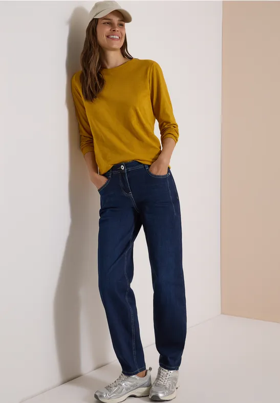Eine lächelnde Frau lehnt an einer Wand, trägt gelbes Shirt, Jeans und weiße Sneakers.
