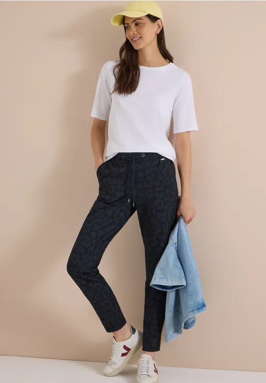 Frau trägt weisses T-Shirt, gemusterte Dunkelhose, weisse Sneaker, gelbe Kappe, hält Jeansjacke.