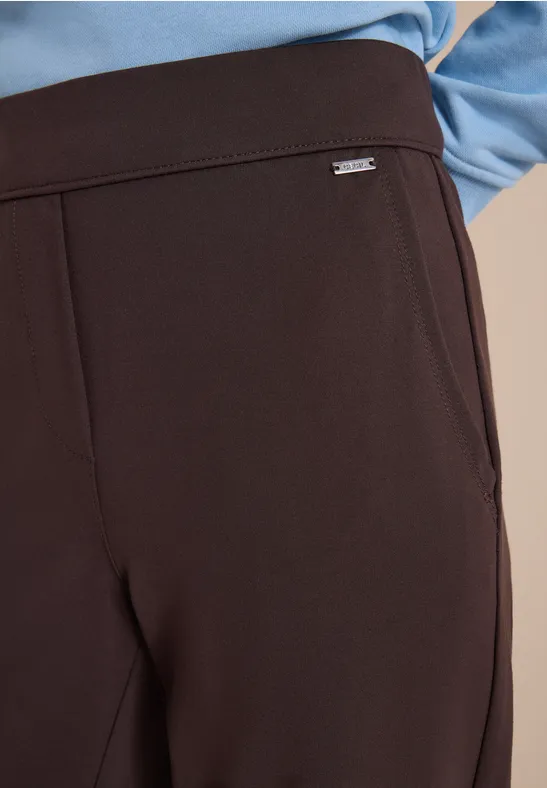 Gros plan sur un pantalon marron foncé élégant avec un petit logo argenté à la taille, porté avec un haut bleu.