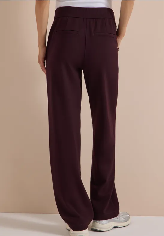 Vue de dos d’une personne portant un pantalon large bordeaux à taille élastique et des baskets claires sur fond beige.